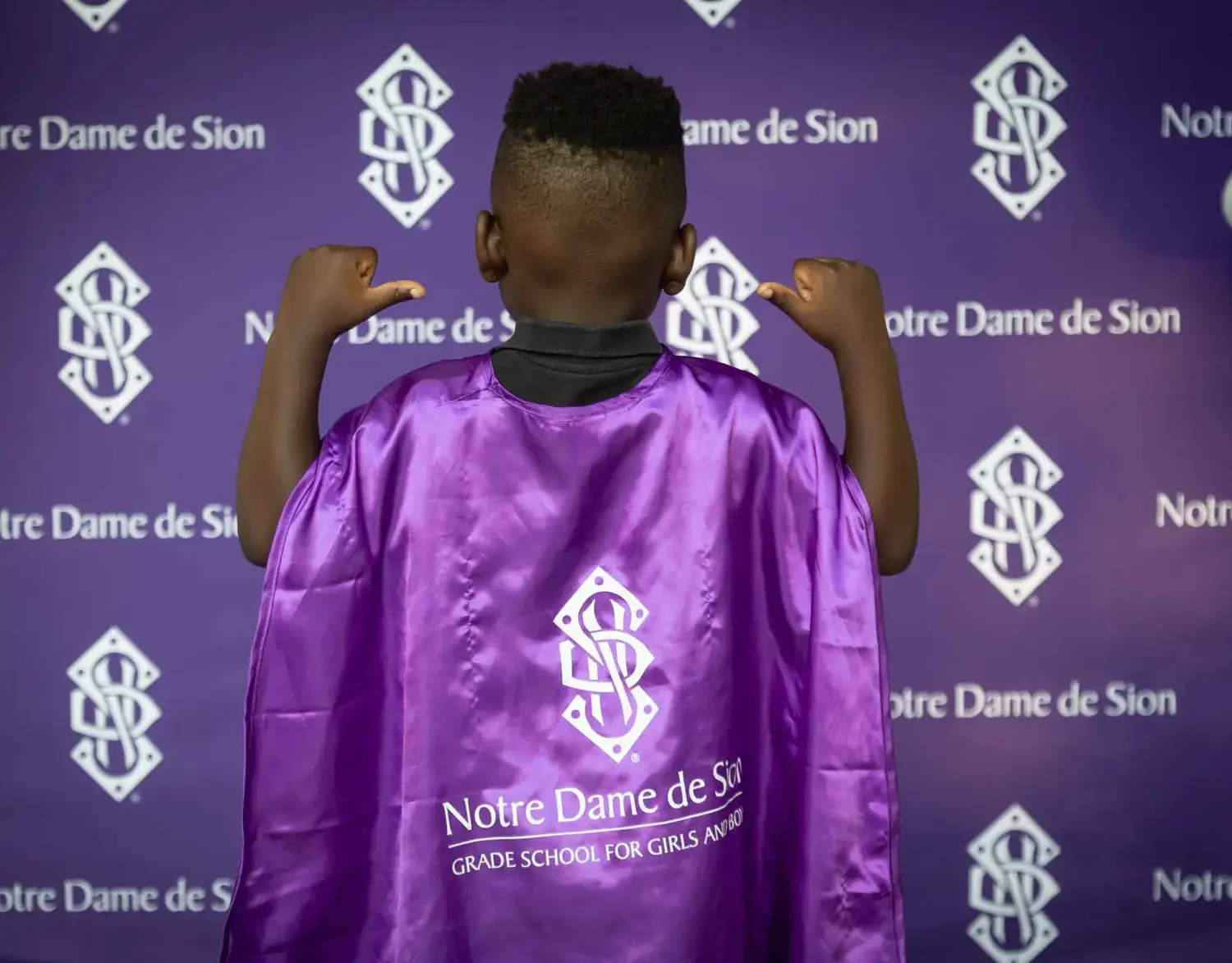 Notre Dame de Sion Grade School student wearing cape and pointing to Sion logo printed on the back.