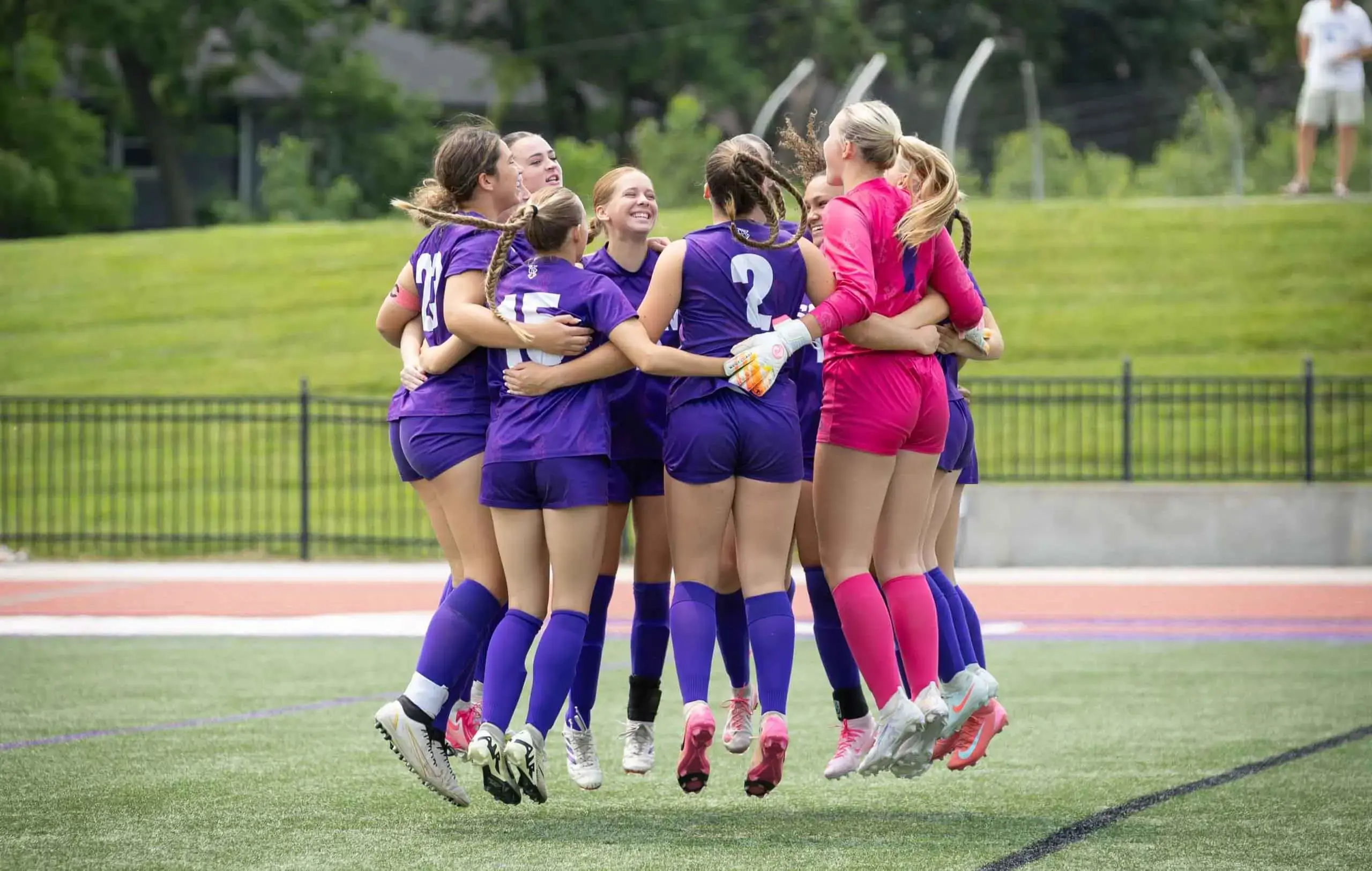 Notre Dame de Sion High School student field hockey team jumping for joy together in a huddle on the pitch.