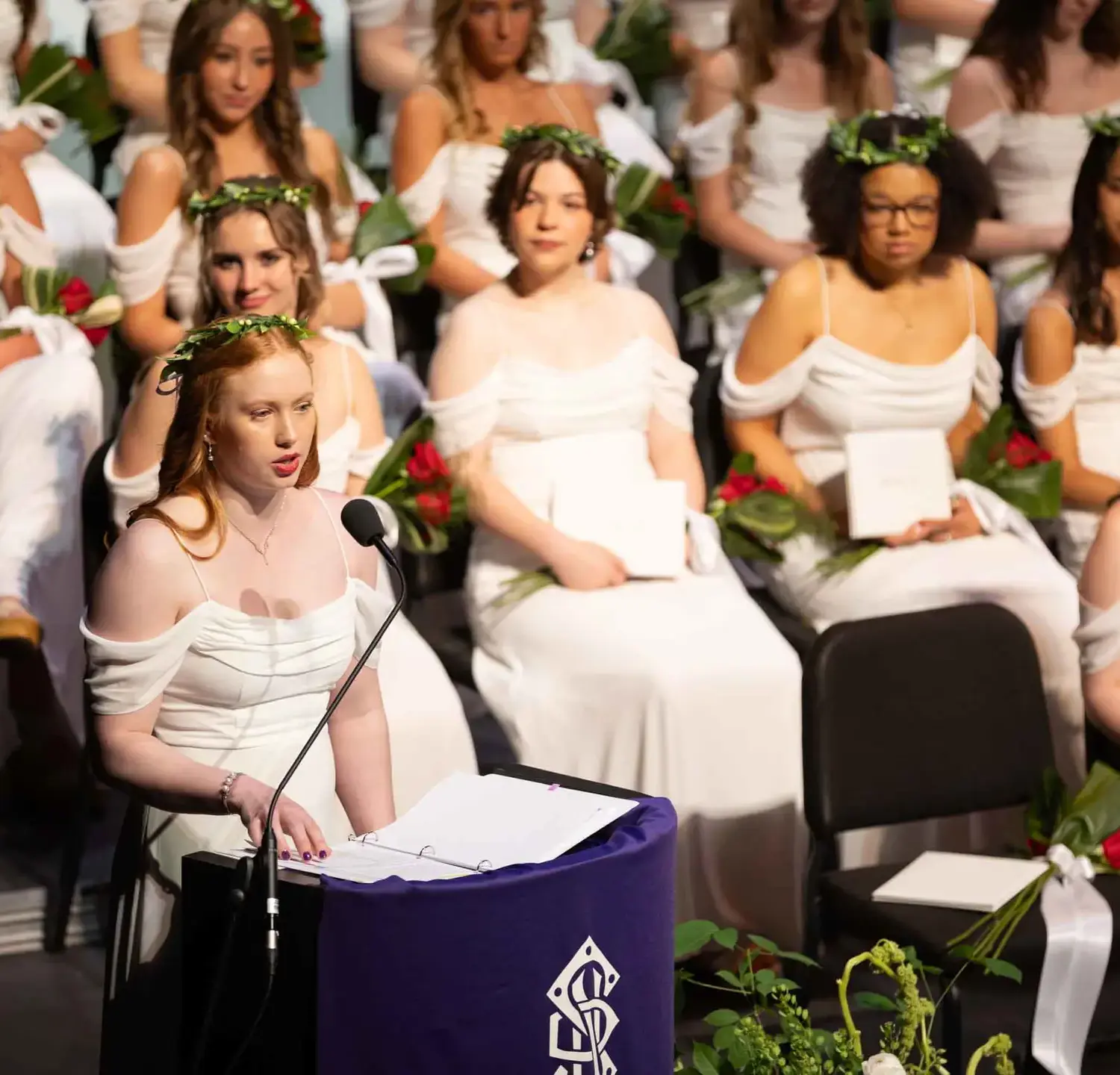 Notre Dame de Sion High School students at graduation, wearing white dresses, listening to peer give speech at lectern.